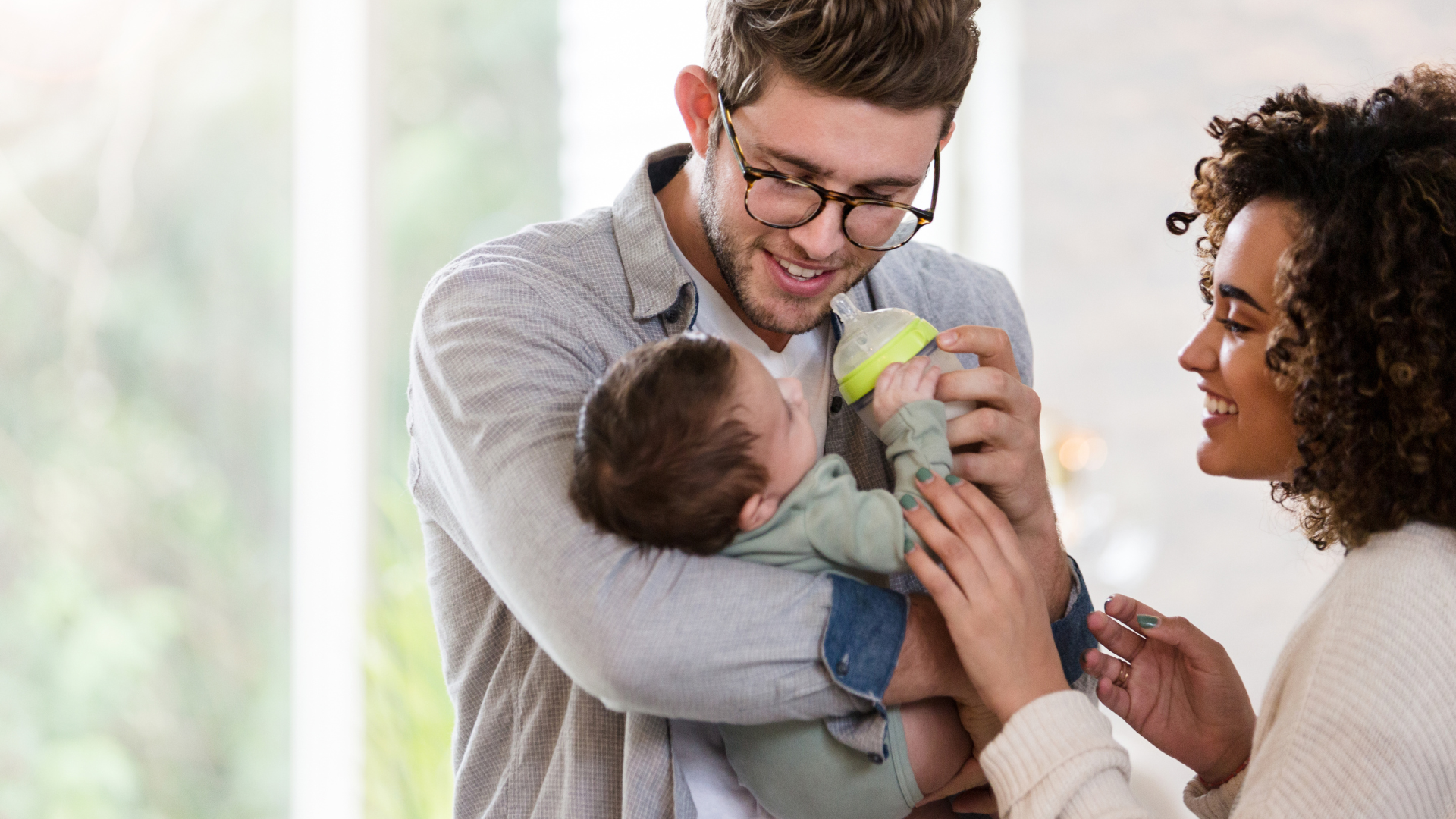 Dad holding baby in arms, feeding with a milk bottle while mum speaks to baby